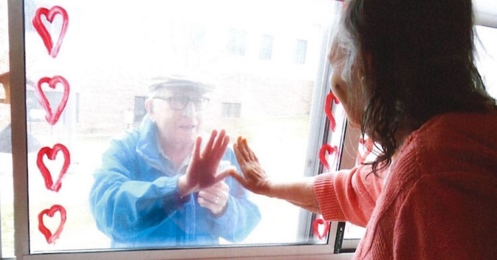 A photograph of two seniors touching opposite sides of a window.